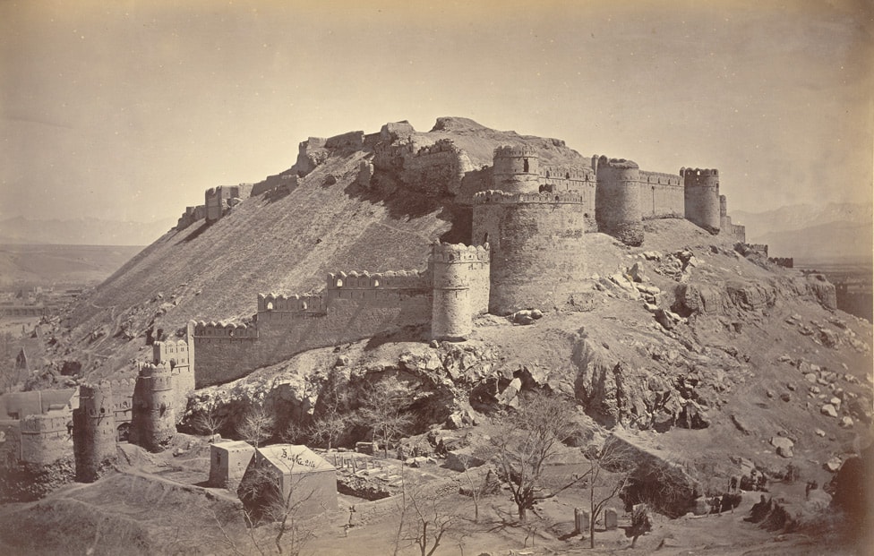 A black-and-white photograph of the upper walls of the Bala Hissar fortress with a cemetery in the foreground.