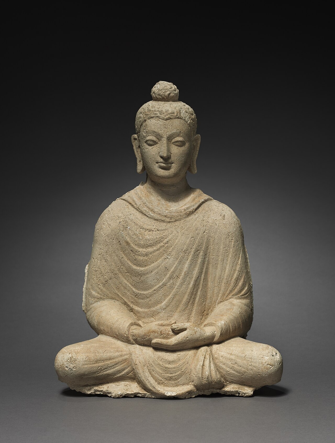A seated Buddha in a robe with hands in his lap, a sculpture from Hadda.