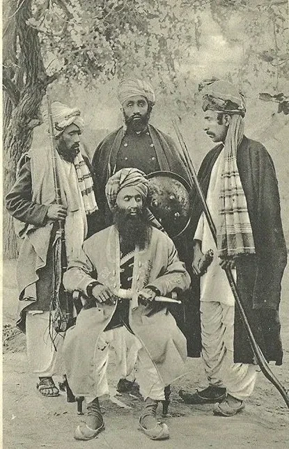Group portrait of an Orakzai Chief and three tribesmen, ca. 1900, , the Chief sits in an armchair wearing a striped turban, an embroidered woolen full-length embroidered coat over a velvet gold-embroidered waistcoat, he wears baggy Pathan pants and traditional leather shoes with curling toes, he carries a Khyber knife in its scabbard with metal locket and chape, the three tribesmen around him are armed with jezail matchlocks, Khyber knife and a metal shield dhal with 4 bosses.