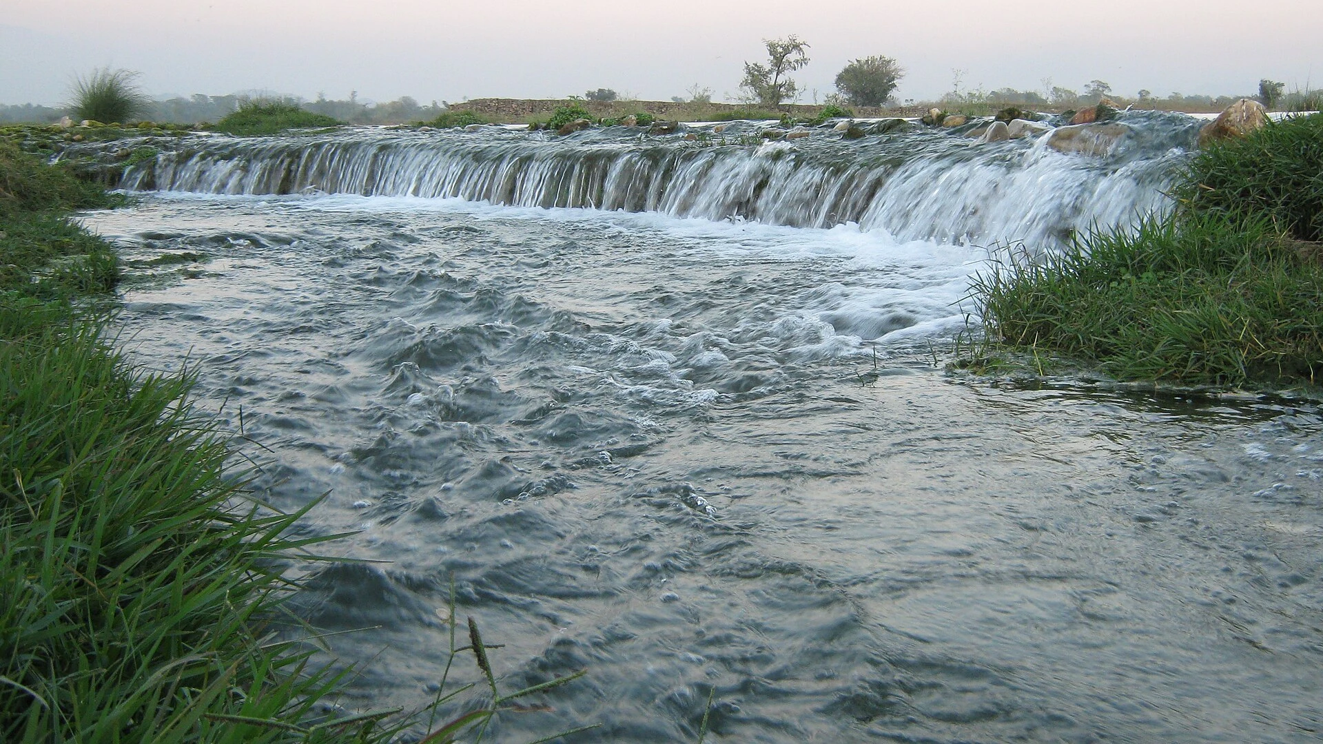 A shallow river crossing flat farmland with hills behind